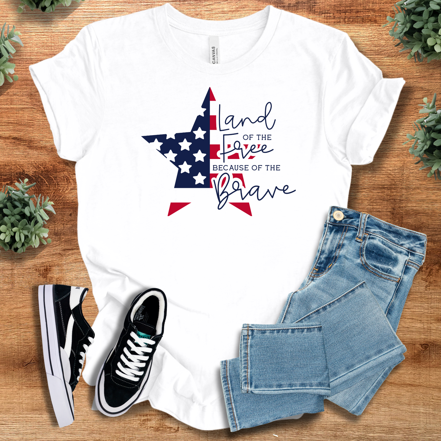 White t-shirt with 'Land Of The Free Because Of The Brave T‑Shirt' text, flat lay styled on a wooden surface.