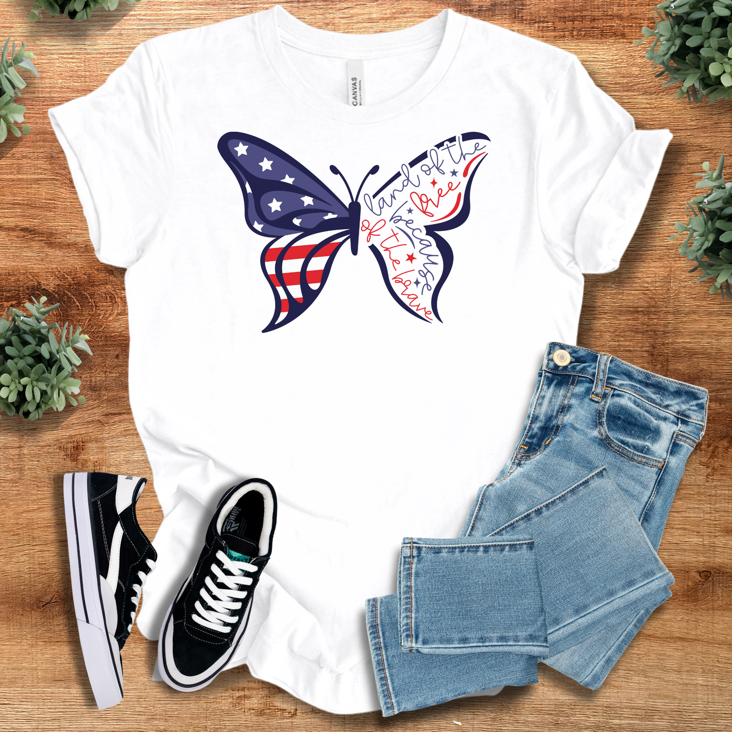 White t-shirt with 'Butterfly Flag Land Of The Free T‑Shirt' text, flat lay styled on a wooden surface.