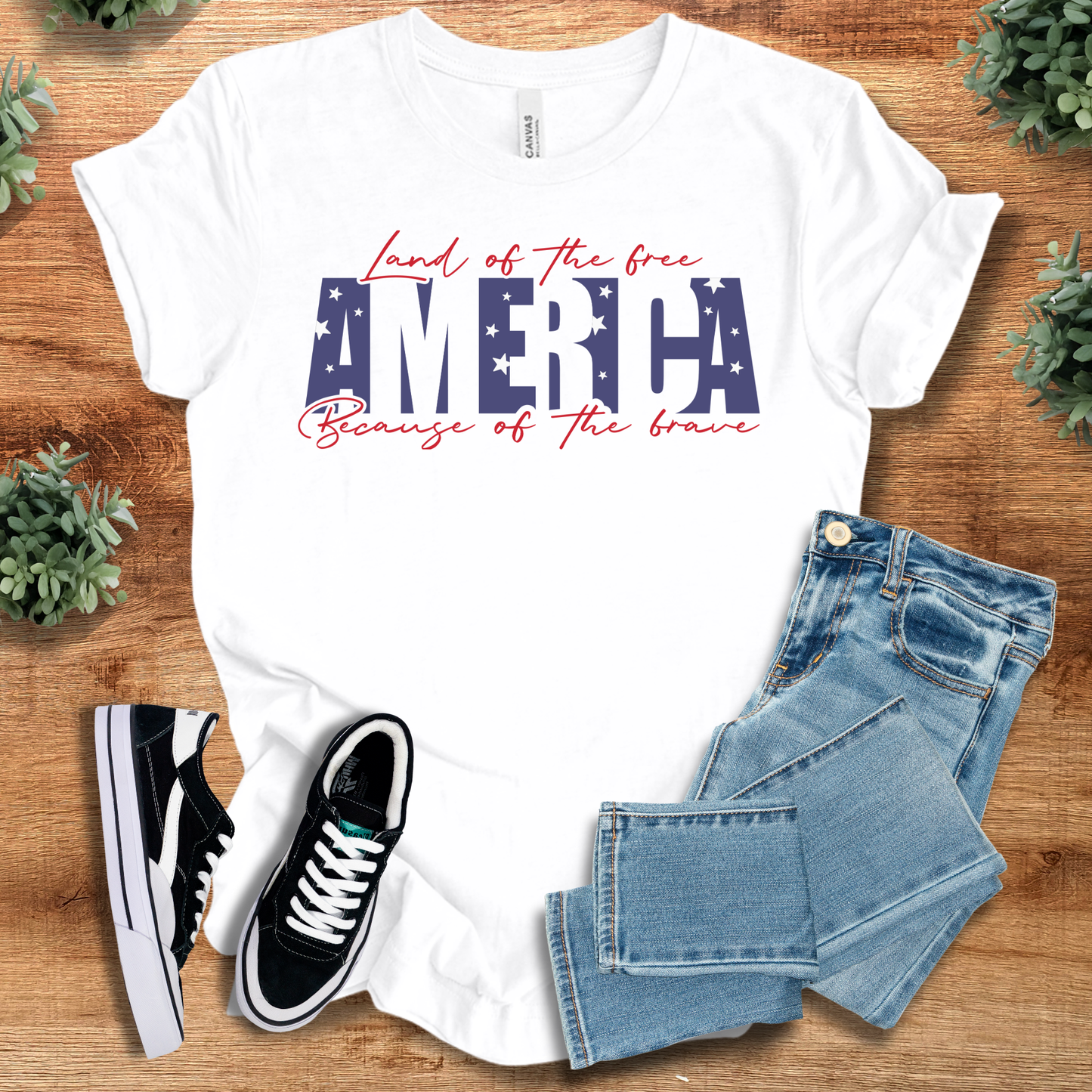 White t-shirt with 'America Land Of The Free T‑Shirt' text, flat lay styled on a wooden surface.