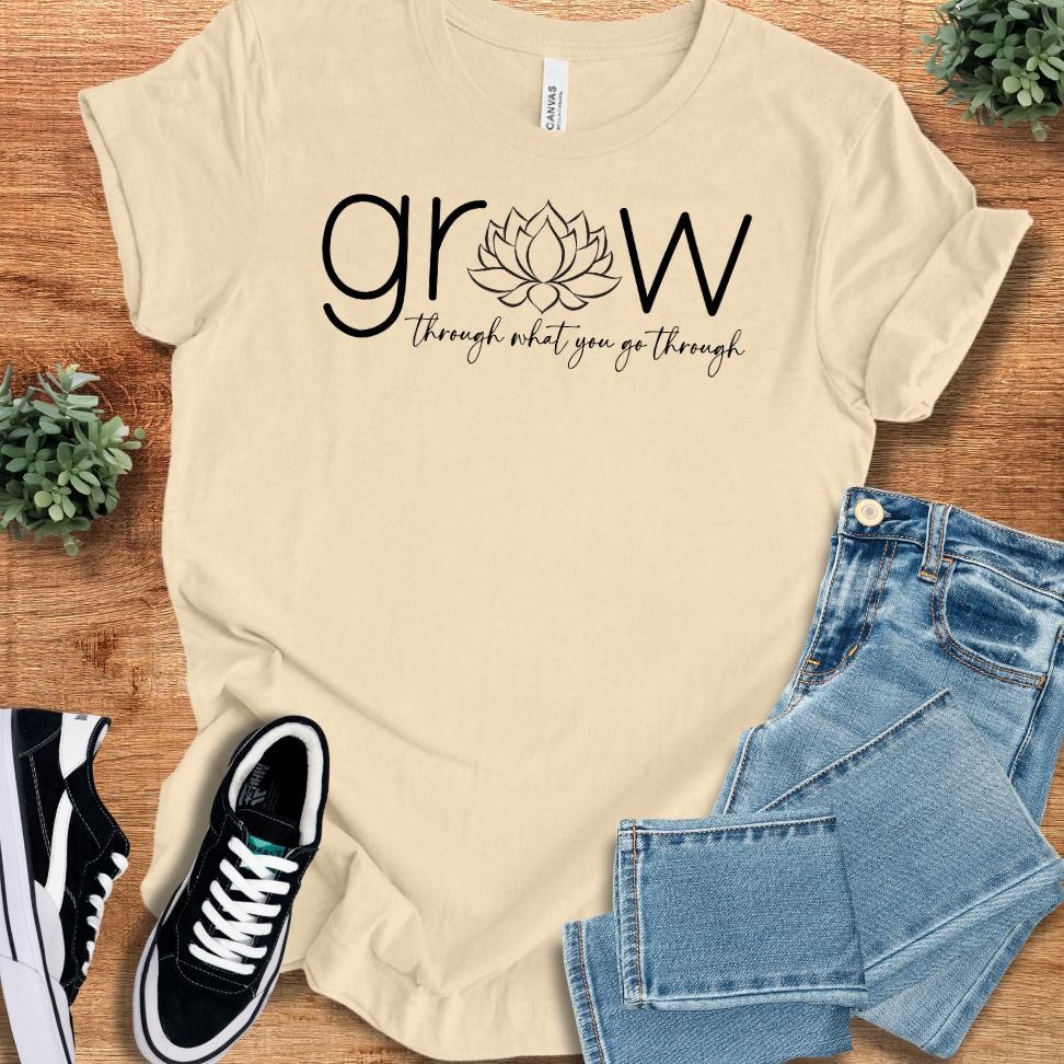 Soft-cream t-shirt with 'Grow Thru What You Go Thru T‑Shirt' text, flat lay styled on a wooden surface.
