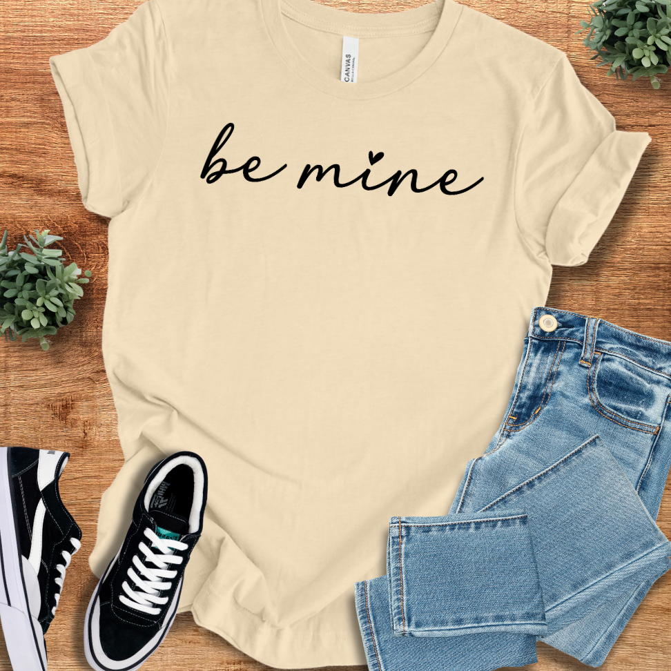 Soft-cream t-shirt with 'Be Mine Classic T‑Shirt' text, flat lay styled on a wooden surface.