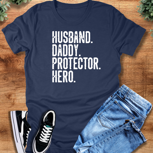Heather-navy t-shirt with 'Husband Daddy Protector Hero T‑Shirt' text, flat lay styled on a wooden surface.