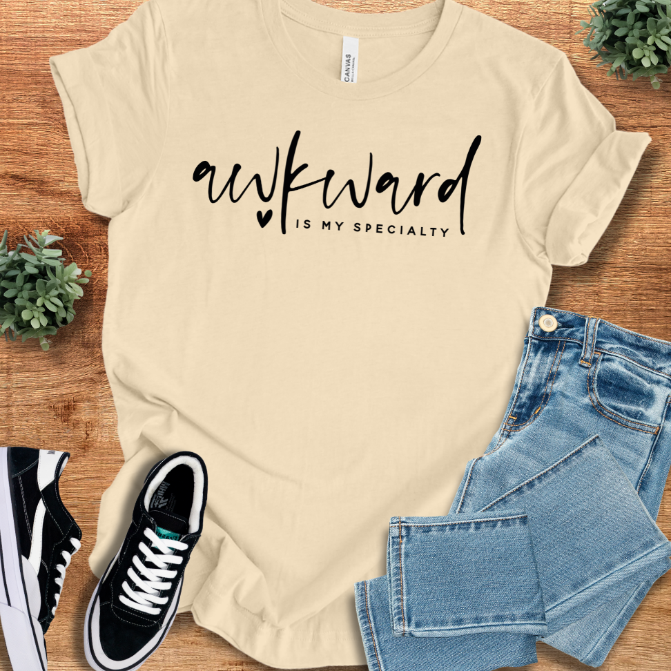Soft-cream t-shirt with 'Awkward T‑Shirt' text, flat lay styled on a wooden surface.