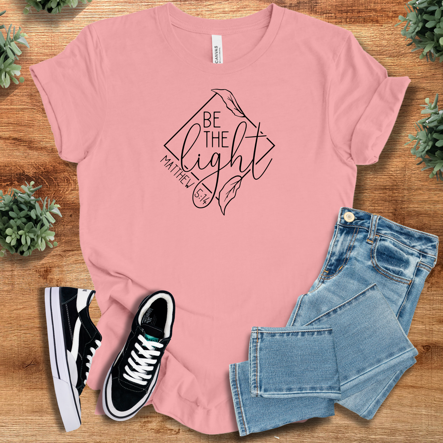 Light pink Be the Light Matthew 5:14 T-shirt with black faith quote and leaf design, styled with jeans for inspirational Christian fashion.