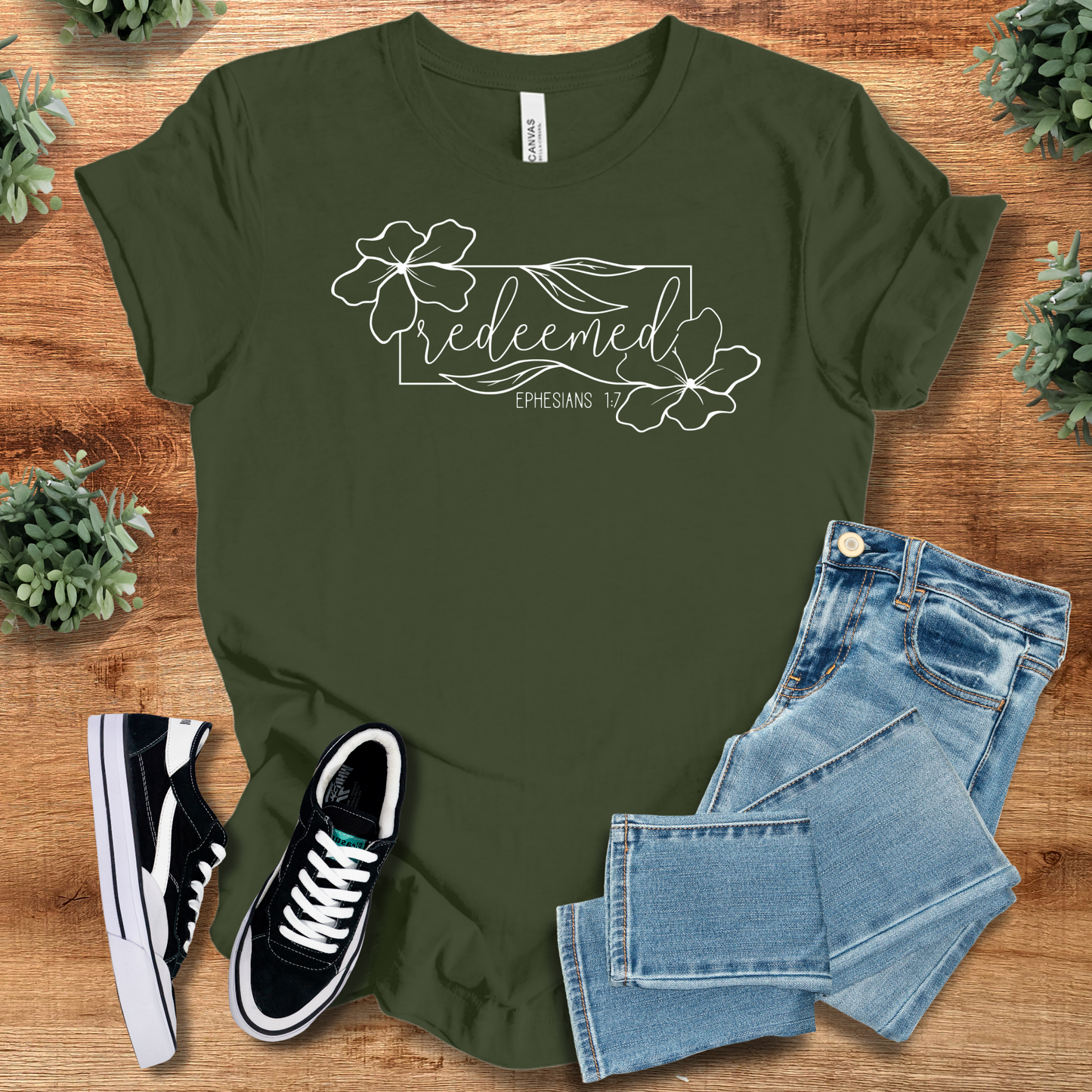 Olive green Redeemed Ephesians 1:7 Tee with floral scripture print, unisex adult fit, styled with jeans and casual shoes on rustic wood.