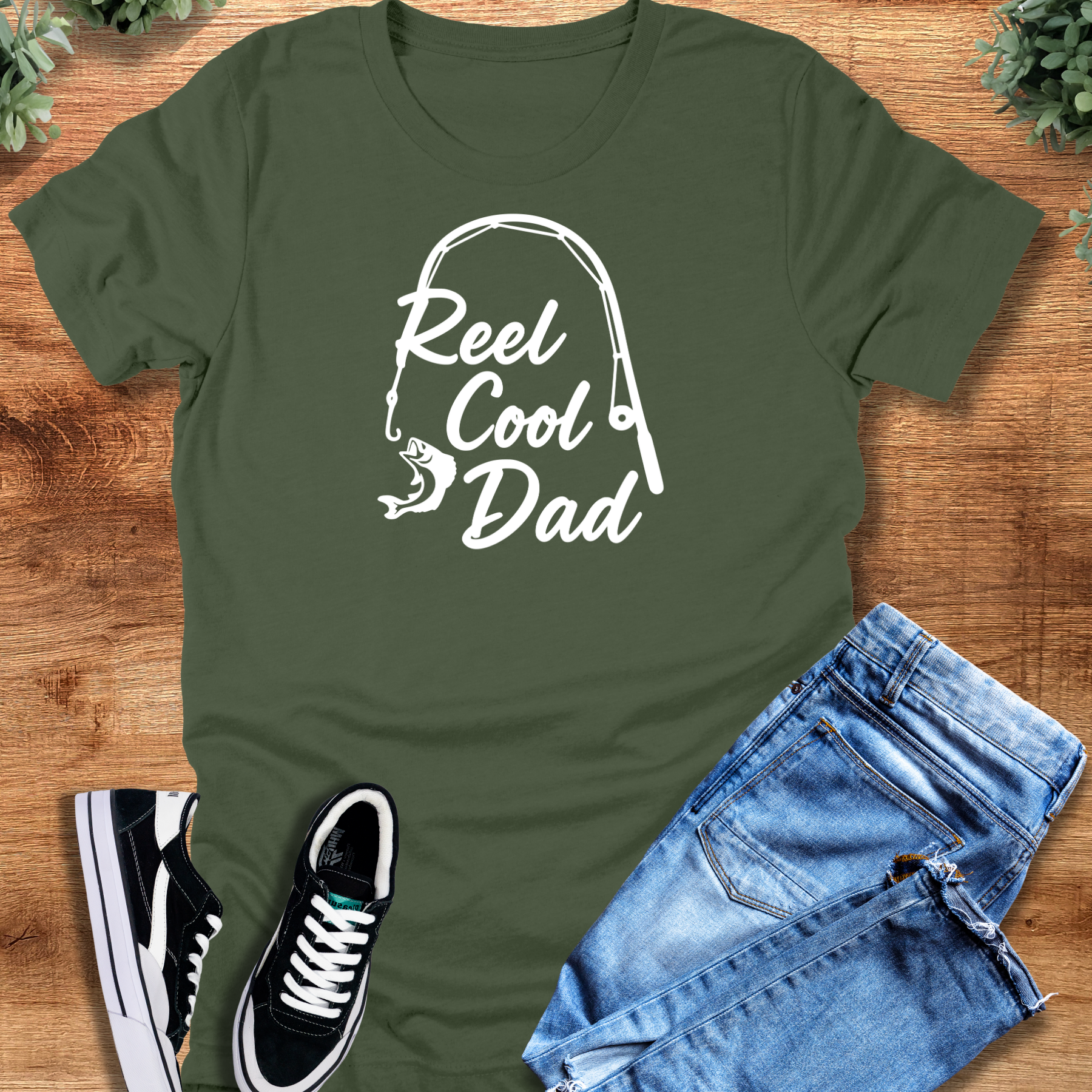 Military-green t-shirt with 'Reel Cool Dad T‑Shirt' text, flat lay styled on a wooden surface.