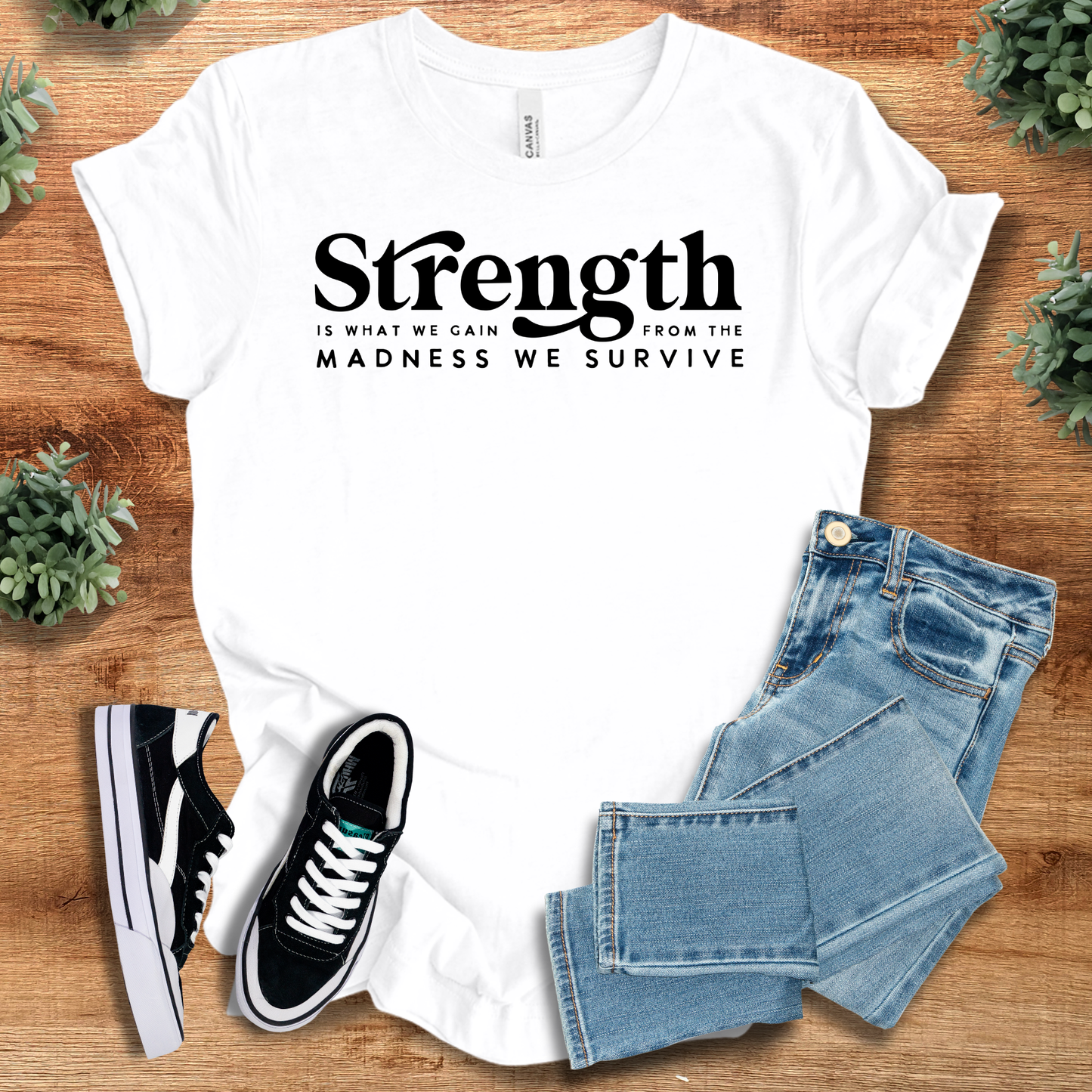 White 'Strength is What We Gain' t-shirt with bold black text, styled with jeans and shoes. Motivational unisex top for everyday empowerment.