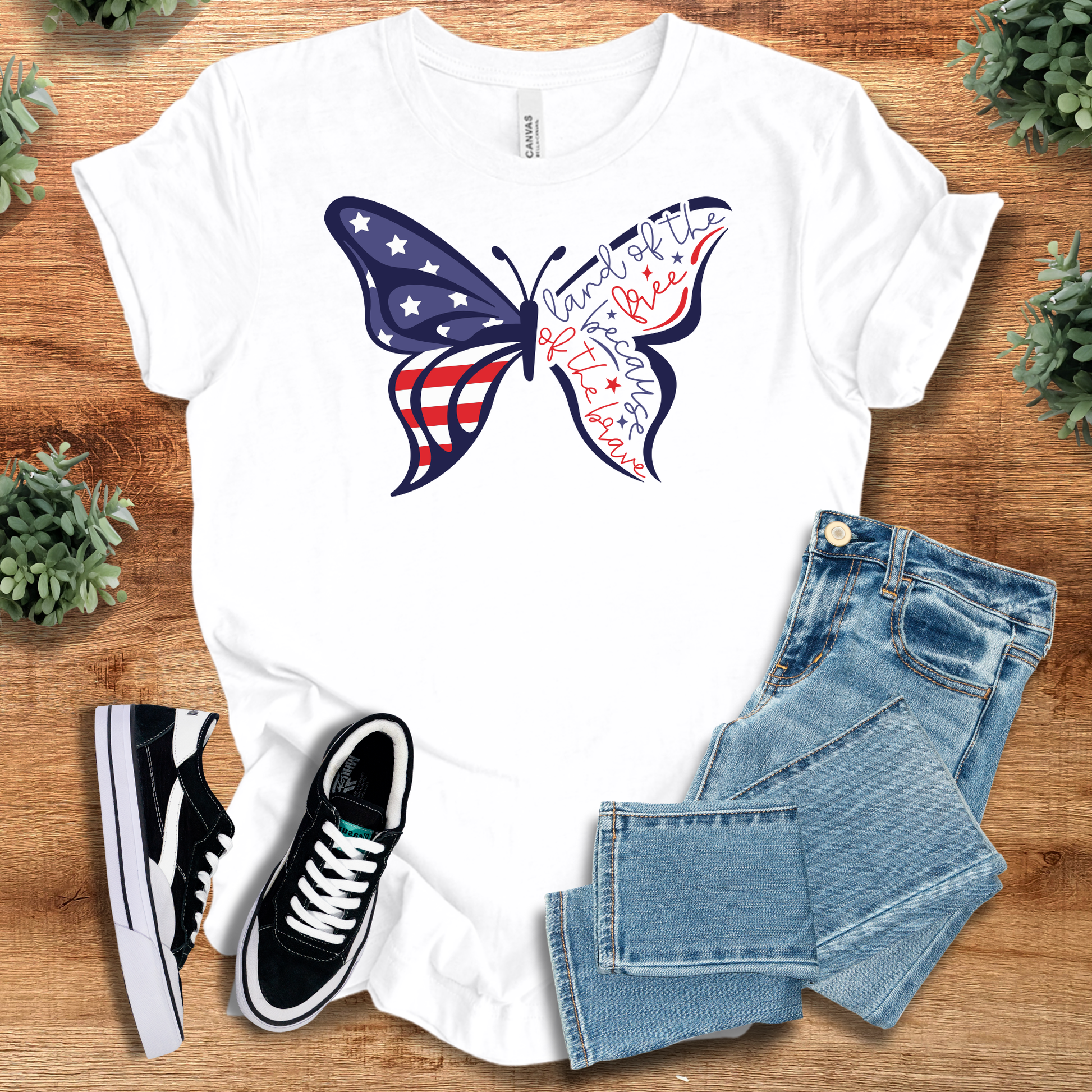 White t-shirt with 'Butterfly Flag Land Of The Free T‑Shirt' text, flat lay styled on a wooden surface.