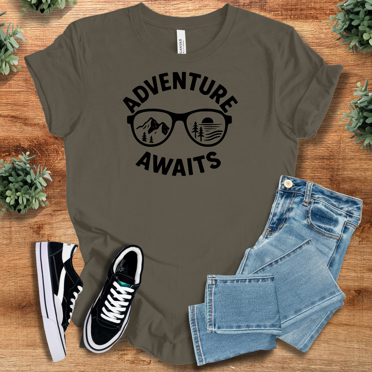 Army t-shirt with 'Adventure Awaits Tee' text, flat lay styled on a wooden surface.