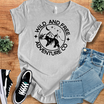 Athletic-heather t-shirt with 'Wild And Free Adventure T‑Shirt' text, flat lay styled on a wooden surface.