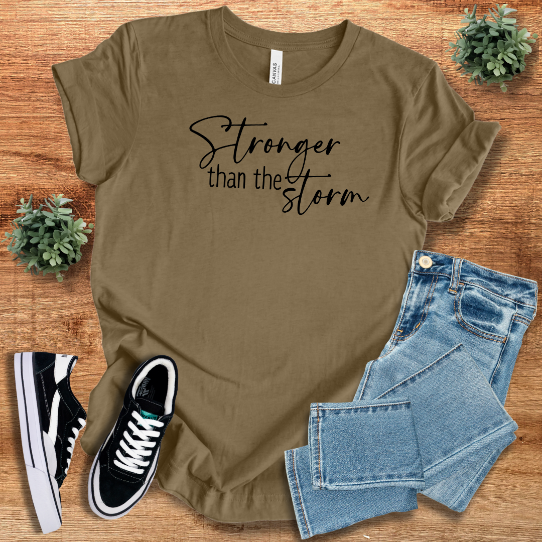Olive green Stronger Than The Storm T-Shirt featuring black script text, paired with jeans and sneakers for a stylish, motivational outfit.