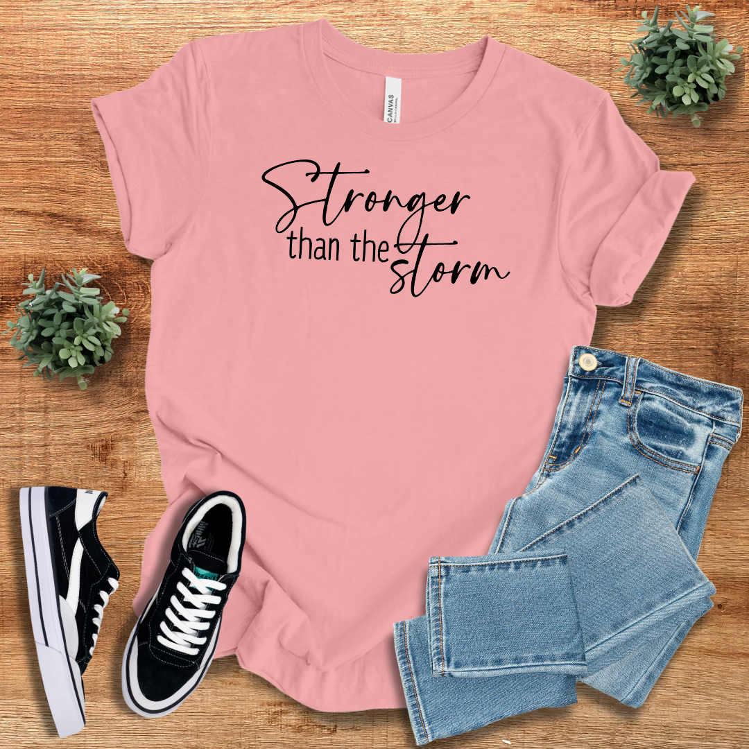 Pink Stronger Than The Storm T-Shirt featuring black script text, paired with jeans and sneakers for a feminine, empowering fashion statement.