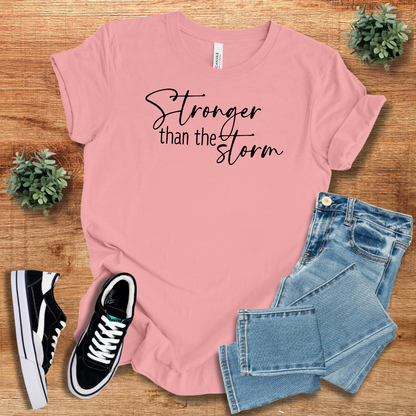 Pink Stronger Than The Storm T-Shirt featuring black script text, paired with jeans and sneakers for a feminine, empowering fashion statement.