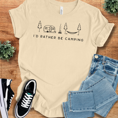 Soft-cream t-shirt with 'I'd Rather Be Camping T‑Shirt' text, flat lay styled on a wooden surface.