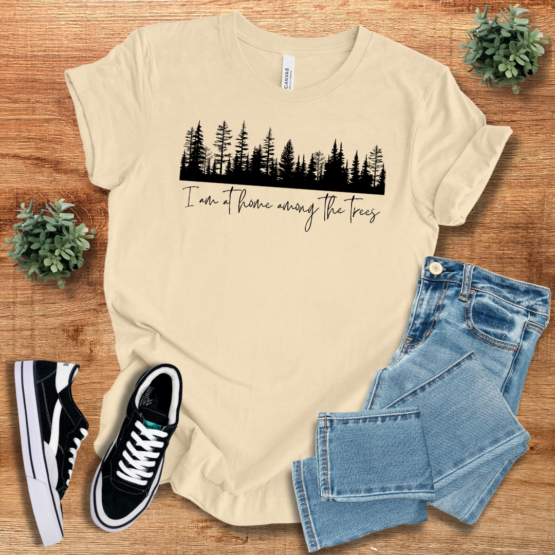 Beige nature lover T-shirt featuring a black tree line design and 'I'm At Home Among The Trees' text. Comfortable and stylish for outdoor adventures.
