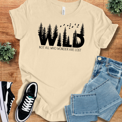 Soft-cream t-shirt with 'Wild, Not All Who T‑Shirt' text, flat lay styled on a wooden surface.