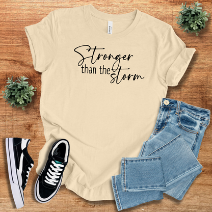 Beige Stronger Than The Storm T-Shirt with black script text, styled with denim jeans and sneakers for a neutral, resilient fashion statement.