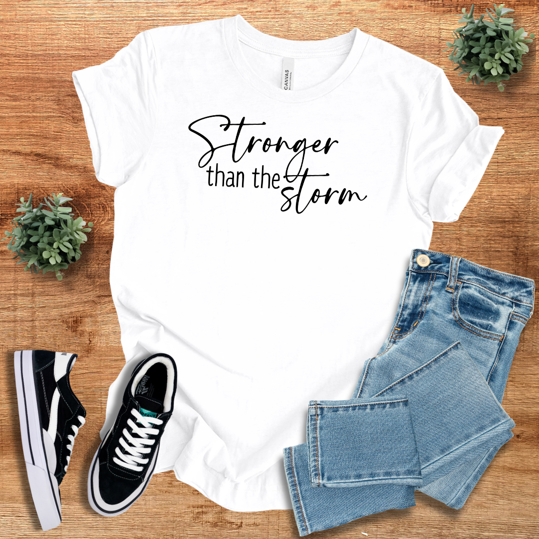 White Stronger Than The Storm T-Shirt with black script text, styled with denim jeans and sneakers for a clean, empowering everyday look.