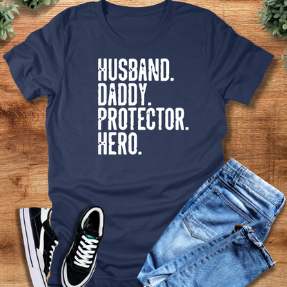 Heather-navy t-shirt with 'Husband Daddy Protector Hero T‑Shirt' text, flat lay styled on a wooden surface.