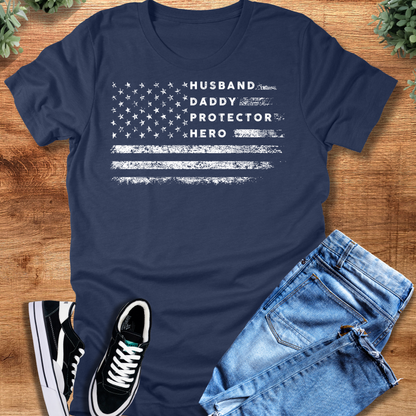Heather-navy t-shirt with 'Husband Daddy Protector Hero Flag T‑Shirt' text, flat lay styled on a wooden surface.