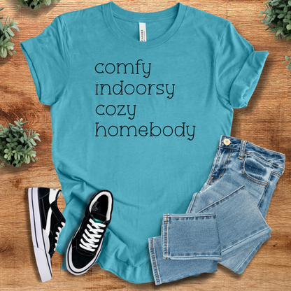 Heather-aqua t-shirt with 'Comfy Indoorsy Cozy Homebody T‑Shirt' text, flat lay styled on a wooden surface.