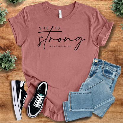Heather-navy t-shirt with 'She Is Strong T‑Shirt' text, flat lay styled on a wooden surface.