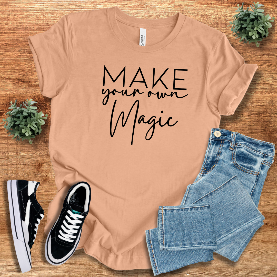 Peach-colored 'Make Your Own Magic' unisex t-shirt with black script text, styled with jeans and sneakers. Soft cotton crew neck tee for casual wear.