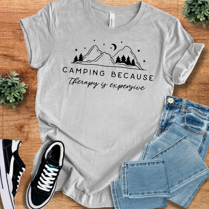 Athletic-heather t-shirt with 'Camping Because Therapy Is Expensive T‑Shirt' text, flat lay styled on a wooden surface.