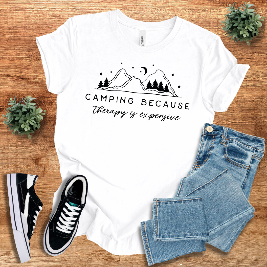 White 'Camping Because Therapy is Expensive' T-shirt with mountain and tree graphic. Comfortable unisex cotton tee for outdoor and casual wear.