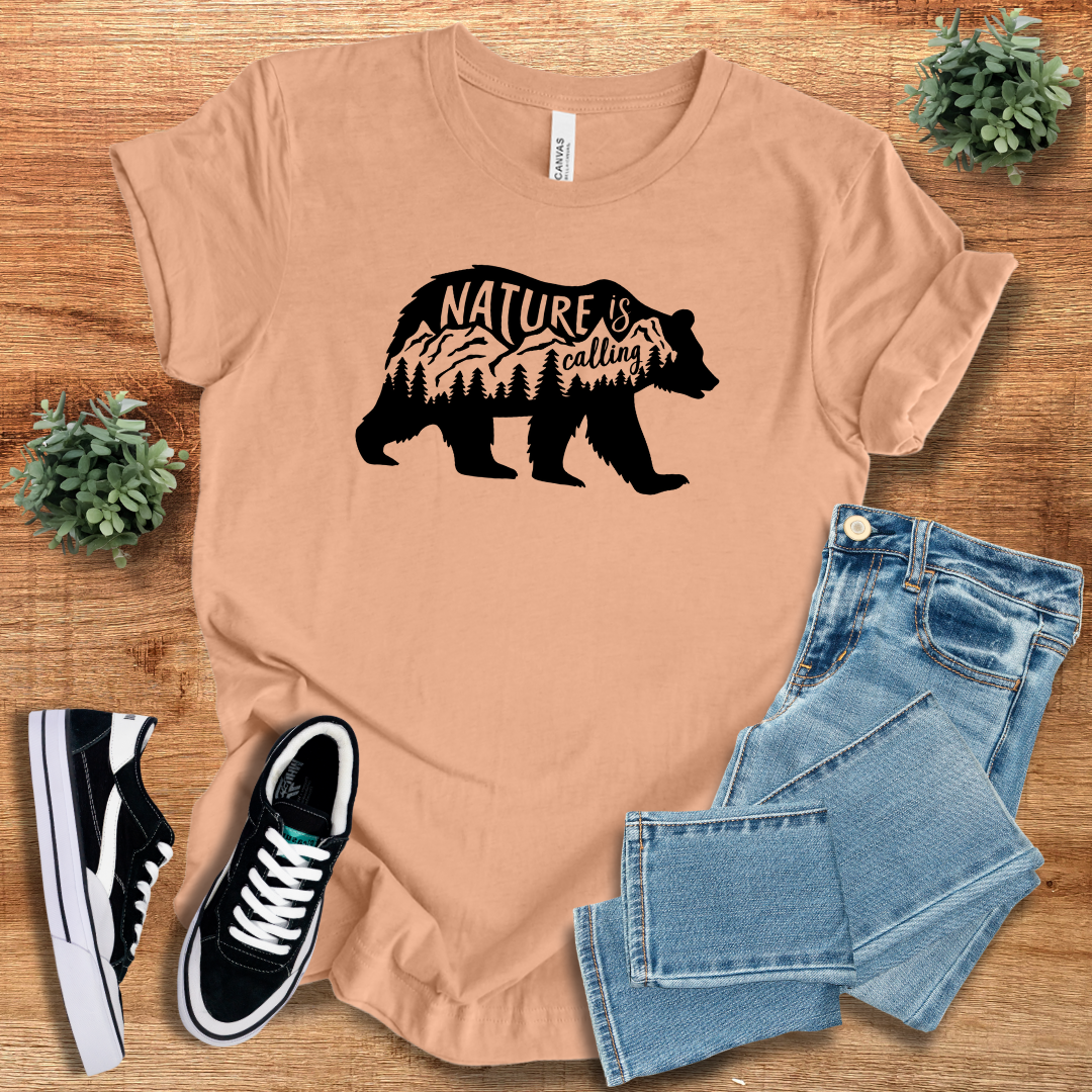 Peach 'Nature Is Calling' t-shirt featuring a black bear silhouette with a mountain and forest design. A breathable and versatile adventure clothing piece.