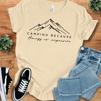 Athletic-heather t-shirt with 'Camping Because Therapy Is Expensive T‑Shirt' text, flat lay styled on a wooden surface.