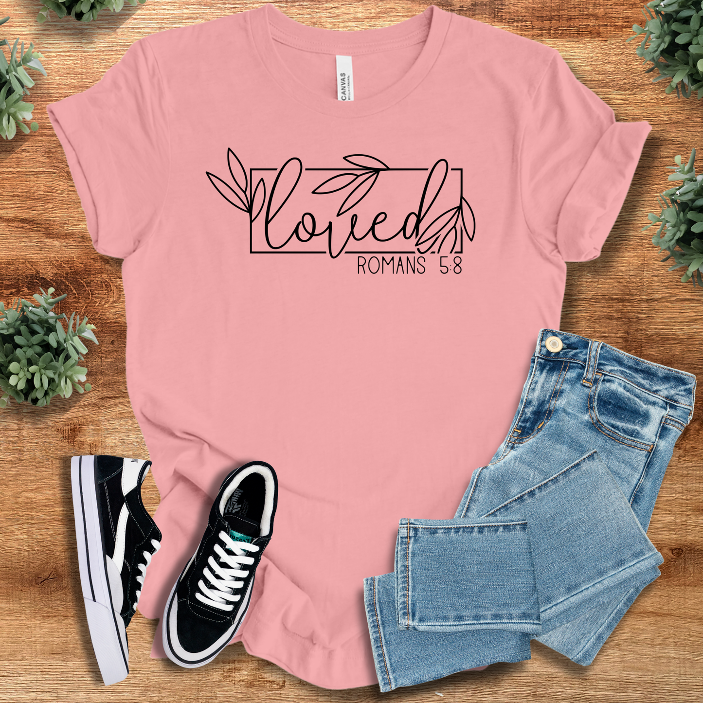 Light pink Loved Romans 5:8 T-Shirt with black script and leaf accents, Christian apparel styled with jeans and casual footwear.