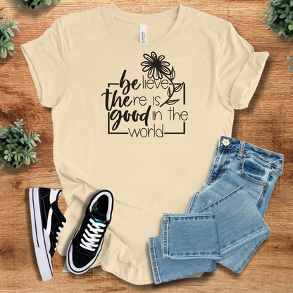Soft-cream t-shirt with 'Believe There Is Good In The World T‑Shirt' text, flat lay styled on a wooden surface.
