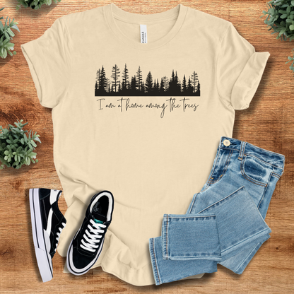 Soft-cream t-shirt with 'I'm At Home Among The Trees T‑Shirt' text, flat lay styled on a wooden surface.