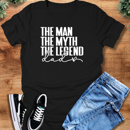 Black t-shirt with 'Dad The Man The Myth The Legend Tee T‑Shirt' text, flat lay styled on a wooden surface.