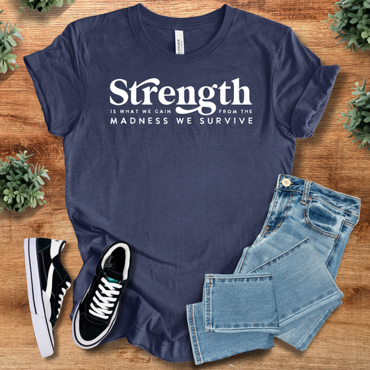 Navy blue 'Strength is What We Gain' tee with bold resilience quote, styled with jeans and sneakers. Unisex motivational shirt for casual wear.