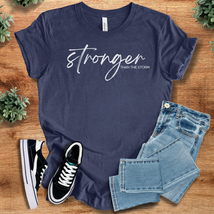 Heather-navy t-shirt with 'Stronger Than The Storm T‑Shirt' text, flat lay styled on a wooden surface.