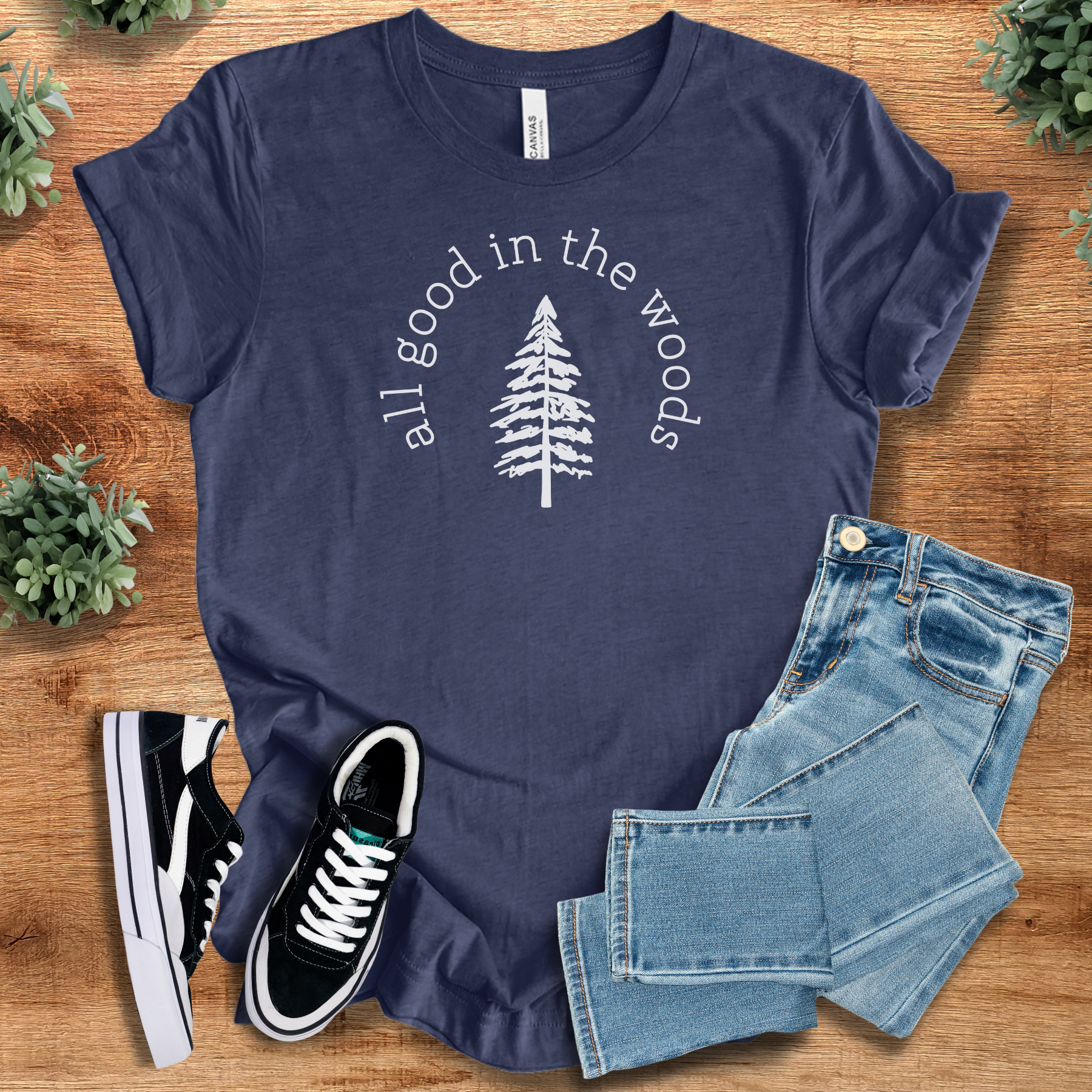 Heather-navy t-shirt with 'All Good In The Woods T‑Shirt' text, flat lay styled on a wooden surface.