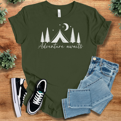Military-green t-shirt with 'Adventure Awaits T‑Shirt' text, flat lay styled on a wooden surface.