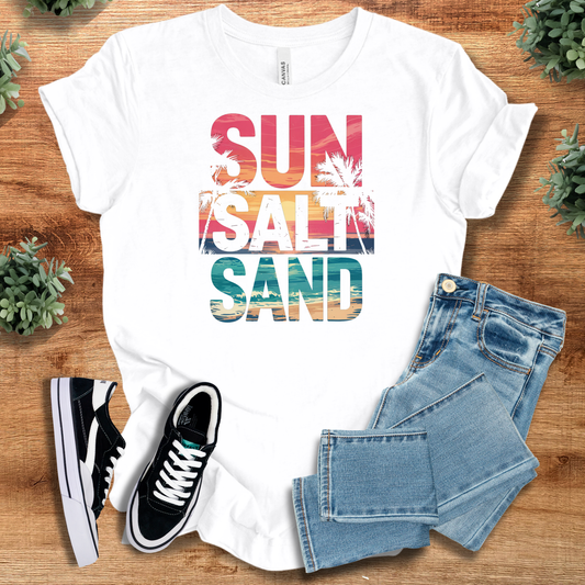 Beach Sun Salt Sand Tee in white with tropical sunset text design, styled with jeans and sneakers. Lightweight 100% cotton beach vacation T-shirt.
